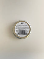 The back of a round metal tin of Geranium & Tomato Leaf nourishing hand cream, with a white label with black text, on a neutral background.