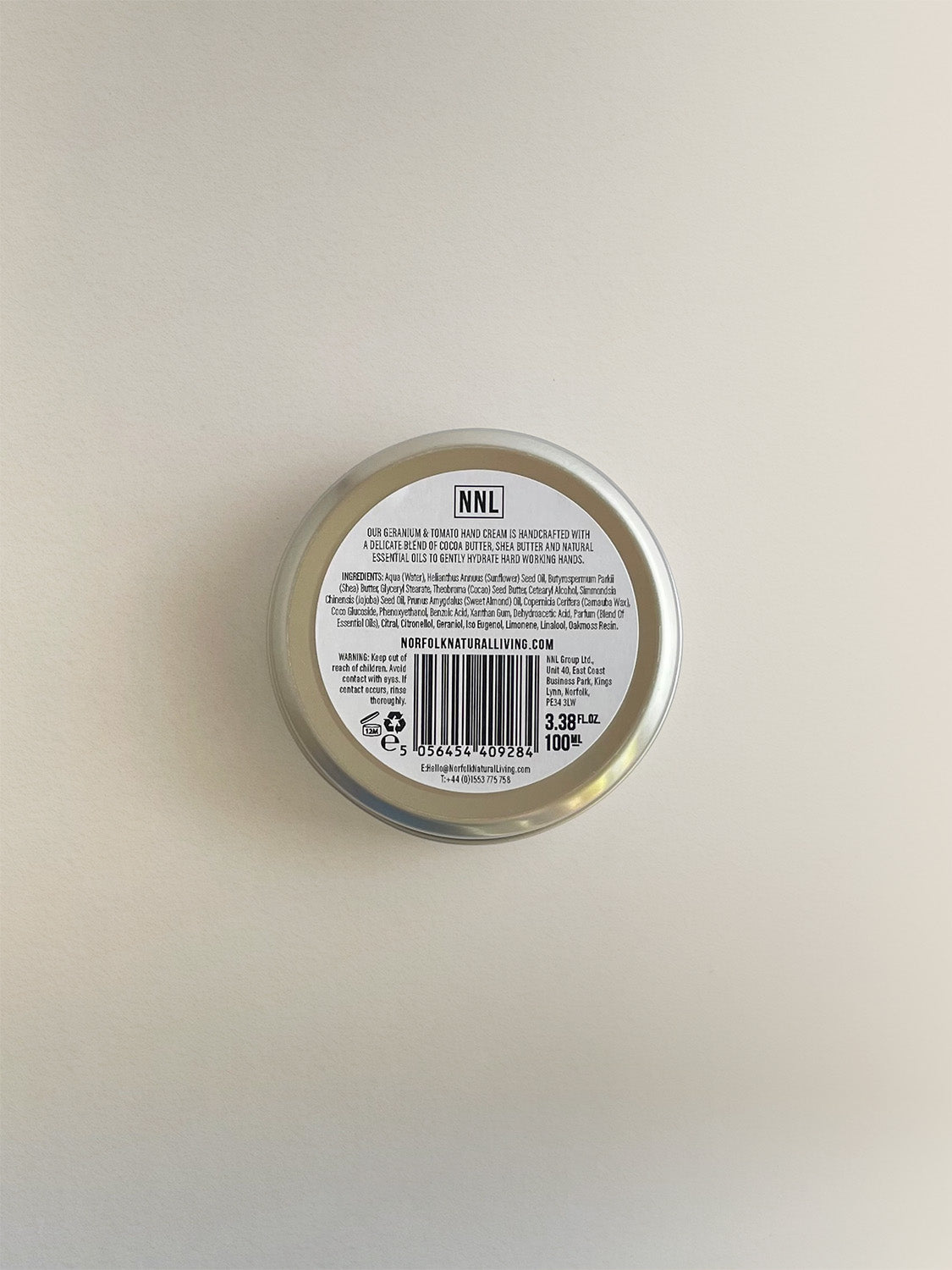 The back of a round metal tin of Geranium & Tomato Leaf nourishing hand cream, with a white label with black text, on a neutral background.