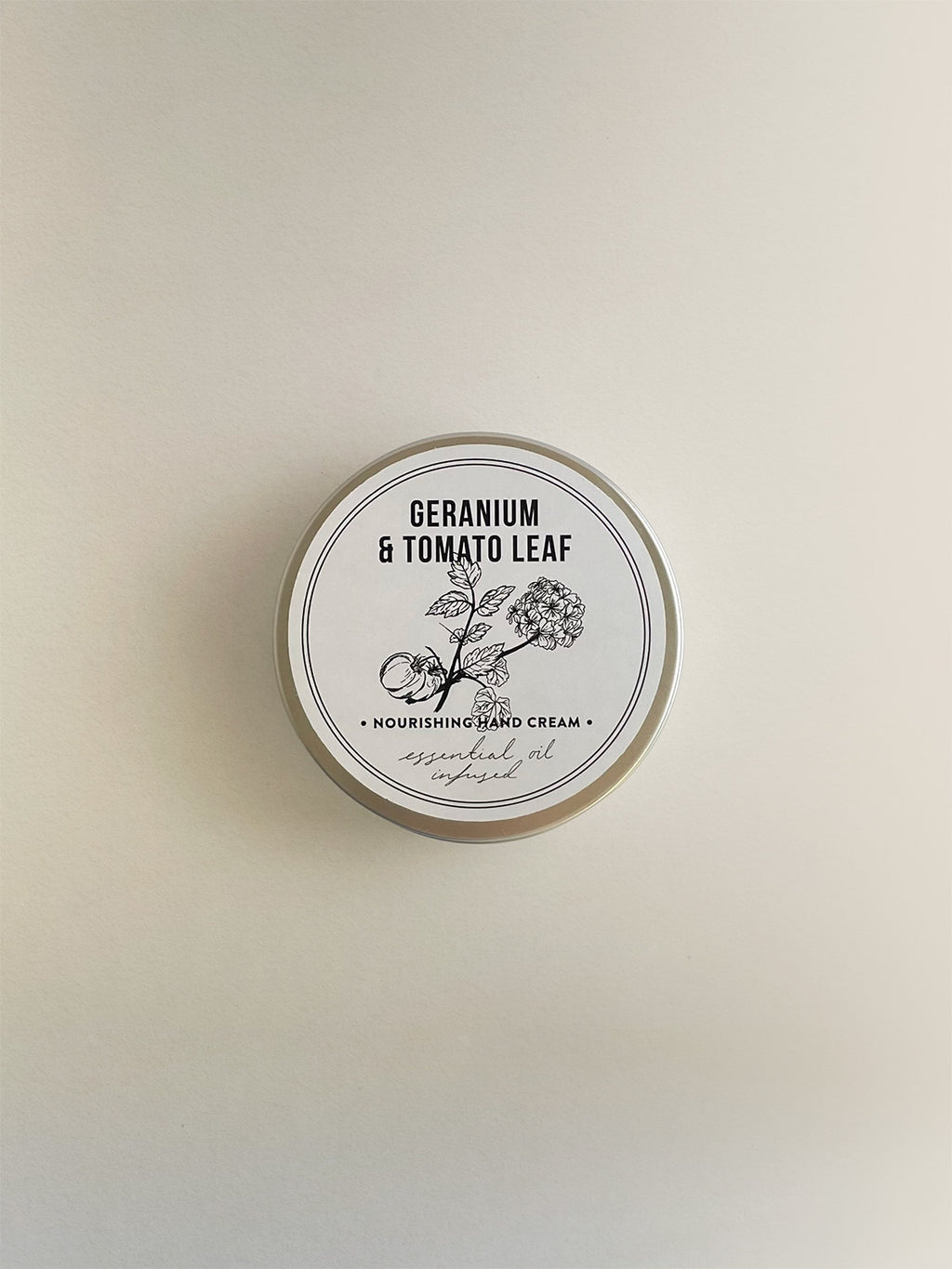 A round metal tin of Geranium & Tomato Leaf nourishing hand cream, with a white label with black botanical illustration, on a neutral background.