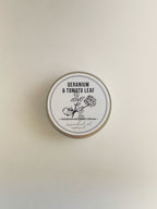 A round metal tin of Geranium & Tomato Leaf nourishing hand cream, with a white label with black botanical illustration, on a neutral background.