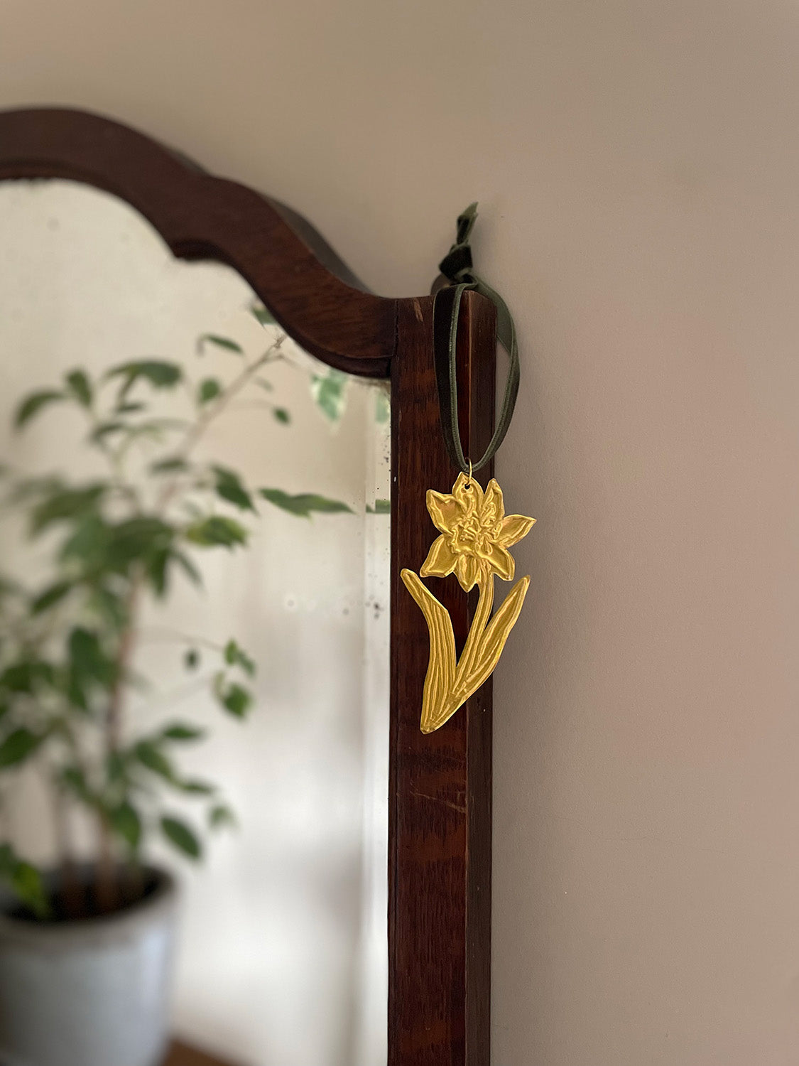 Gold tin daffodil handmade ornament with green velvet ribbon hung on a wooden mirror frame with blurred plant reflection in the background.