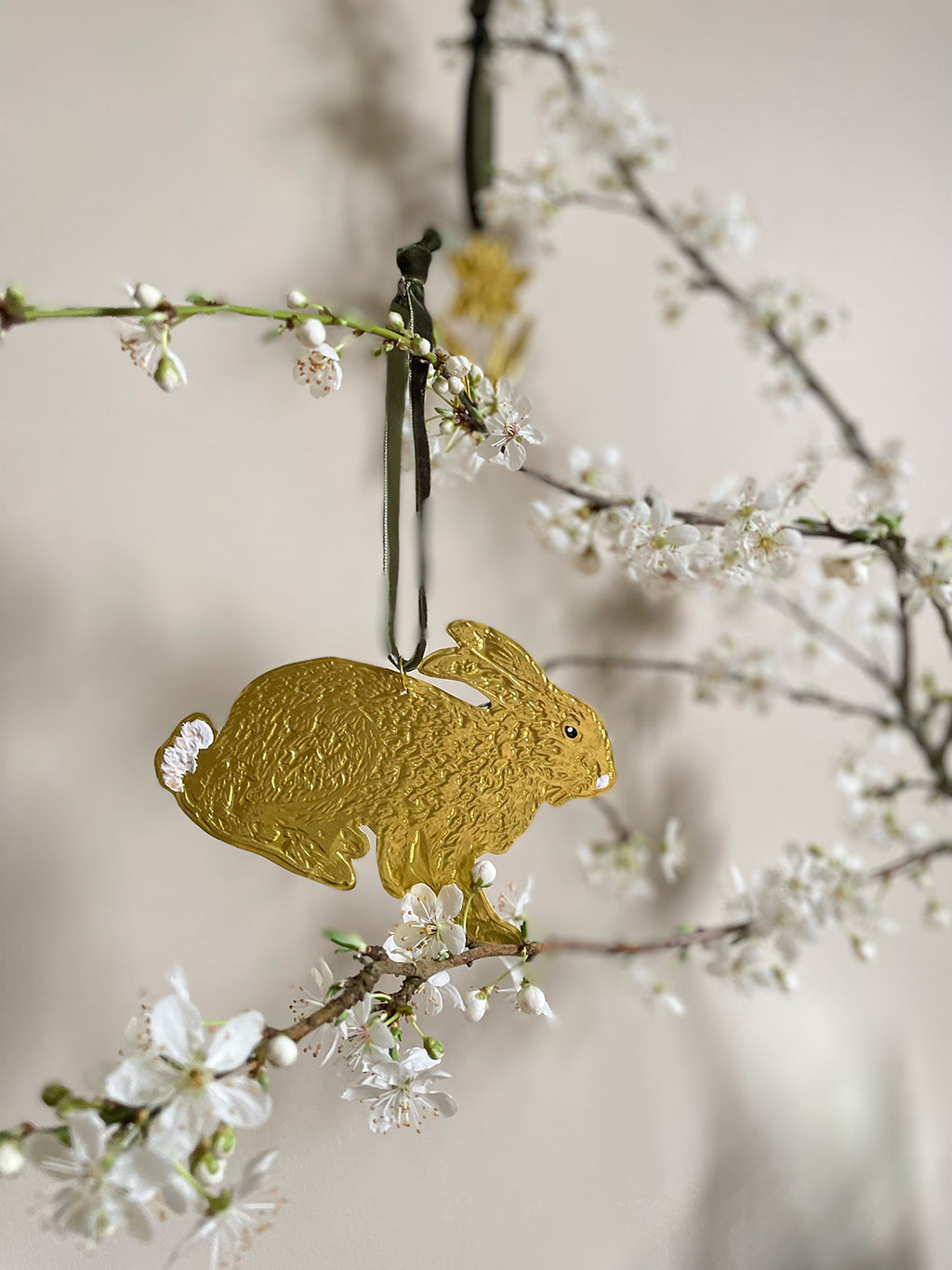 Gold tin hare handmade ornament with green velvet ribbon hung on branches with blossom against a beige background.