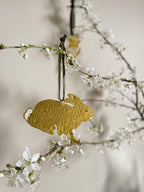Gold tin hare handmade ornament with green velvet ribbon hung on branches with blossom against a beige background.