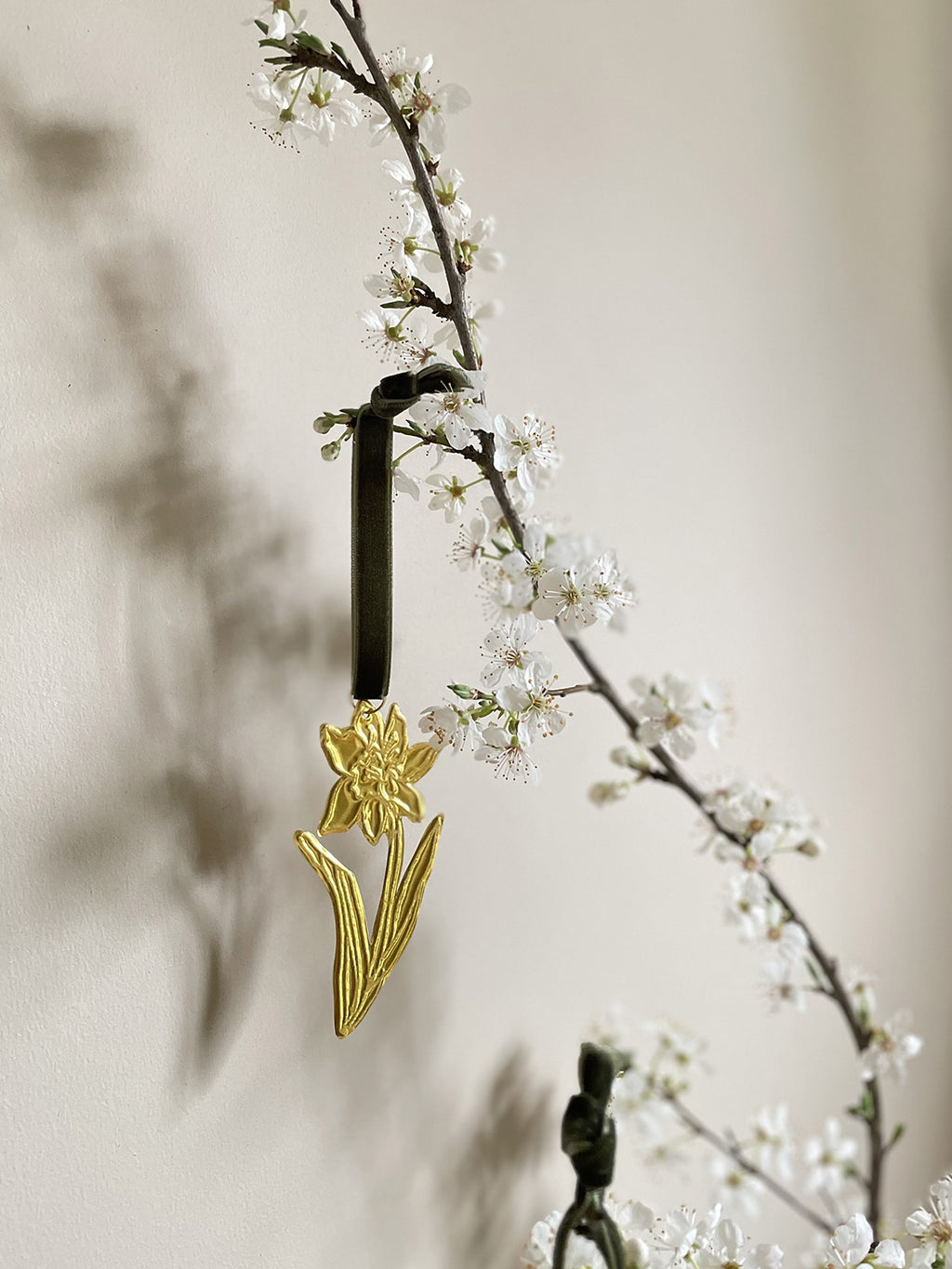 Gold tin daffodil handmade ornament with green velvet ribbon hung on branches with blossom against a beige background.