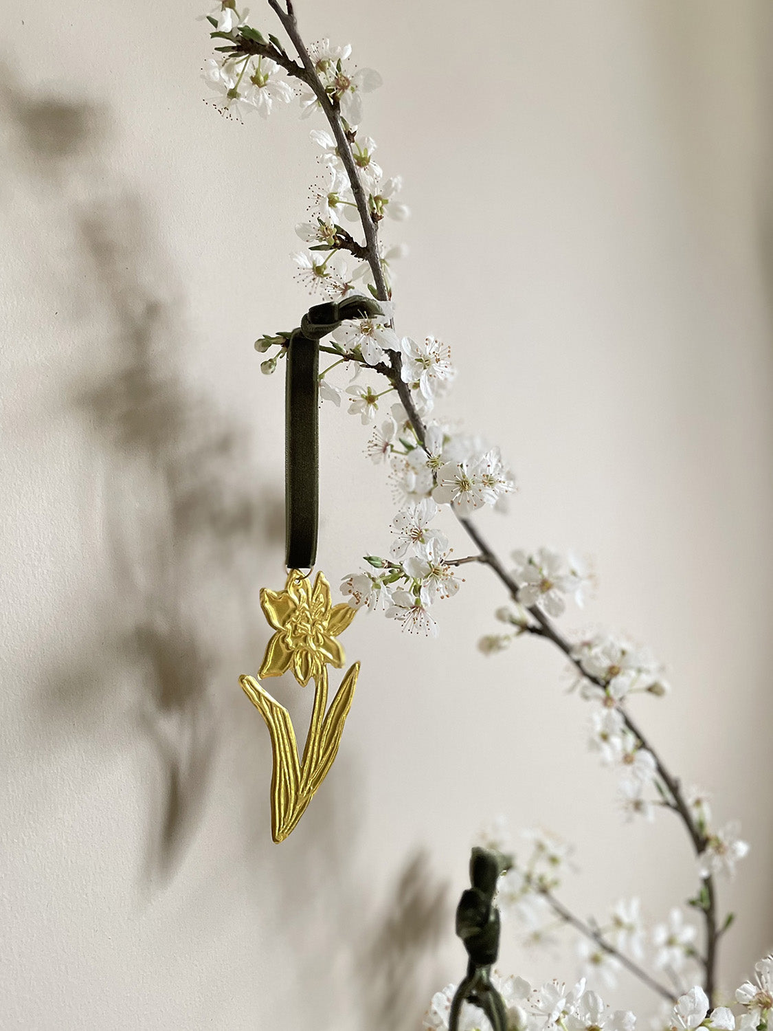 Gold tin daffodil handmade ornament with green velvet ribbon hung on branches with blossom against a beige background.