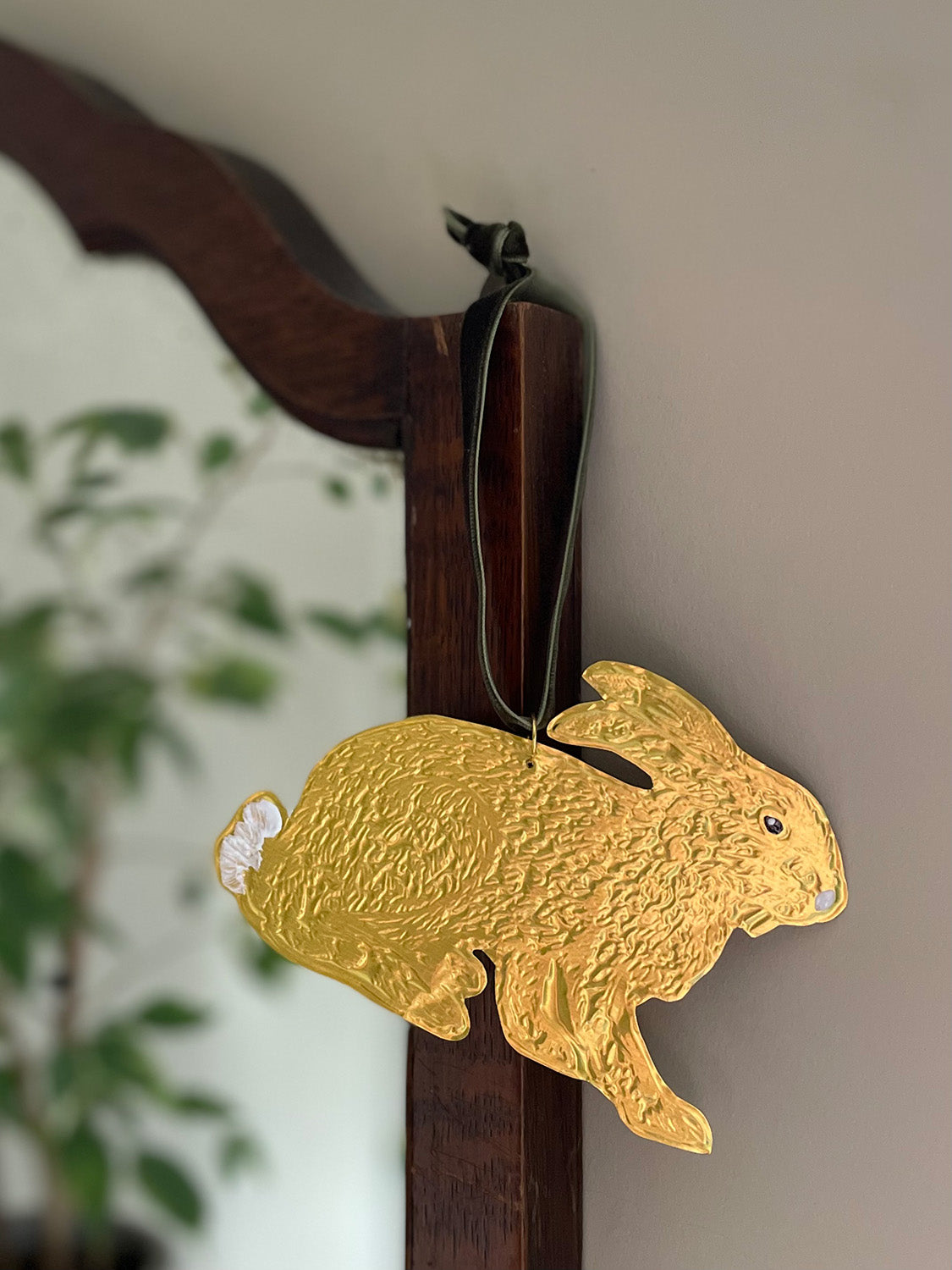 Gold tin hare handmade ornament with green velvet ribbon hung on a wooden mirror frame with blurred plant reflection in the background - close-up.