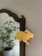 Gold tin hare handmade ornament with green velvet ribbon hung on a wooden mirror frame with blurred plant reflection in the background.