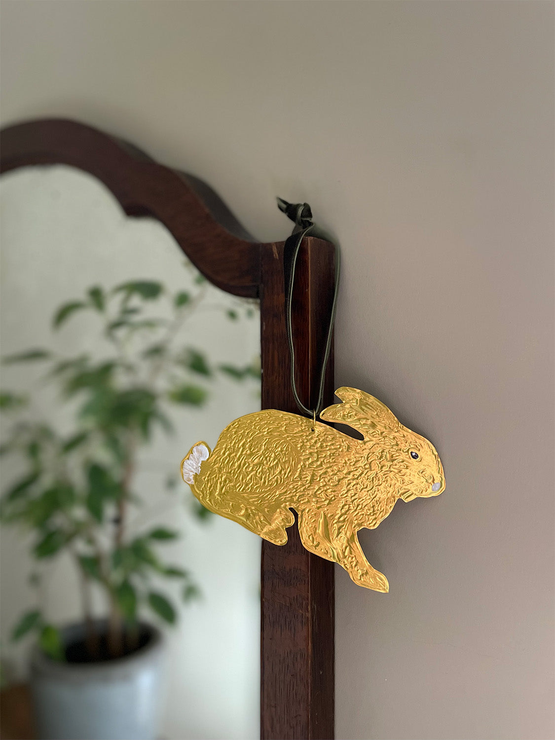 Gold tin hare handmade ornament with green velvet ribbon hung on a wooden mirror frame with blurred plant reflection in the background.