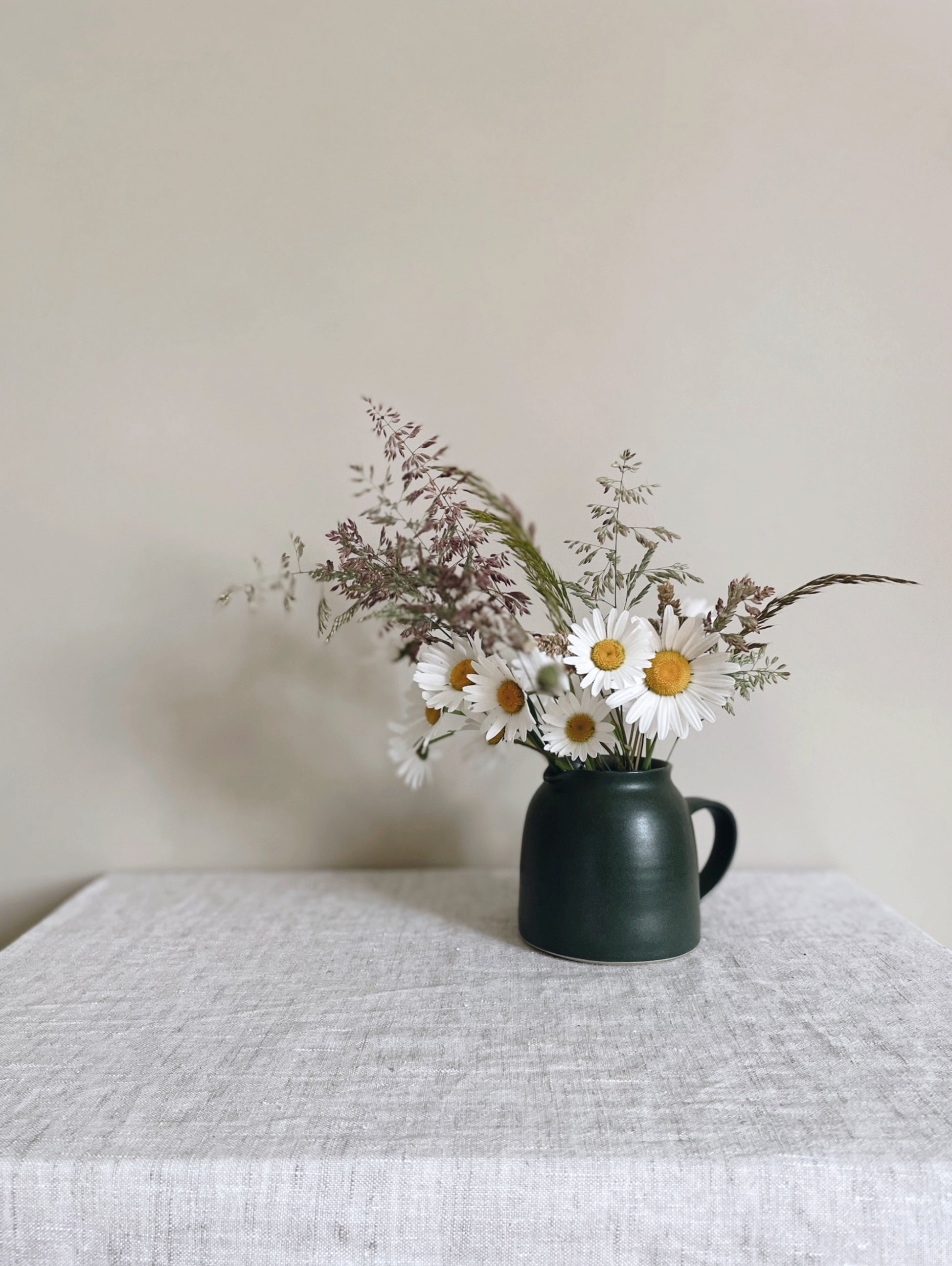 Ceramic jug with handle in deep green glaze with wildflowers, on a beige linen surface with a plain beige wall behind - front view.