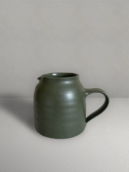 Ceramic jug with handle in deep green glaze on a light taupe surface with a textured taupe wall behind - front view.