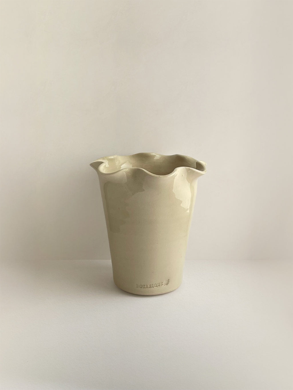 A handmade ceramic cream coloured vase with a frilled, wavy rim and maker's stamp on a neutral background.