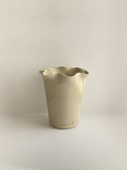 A handmade ceramic cream coloured vase with a frilled, wavy rim and maker's stamp on a neutral background.