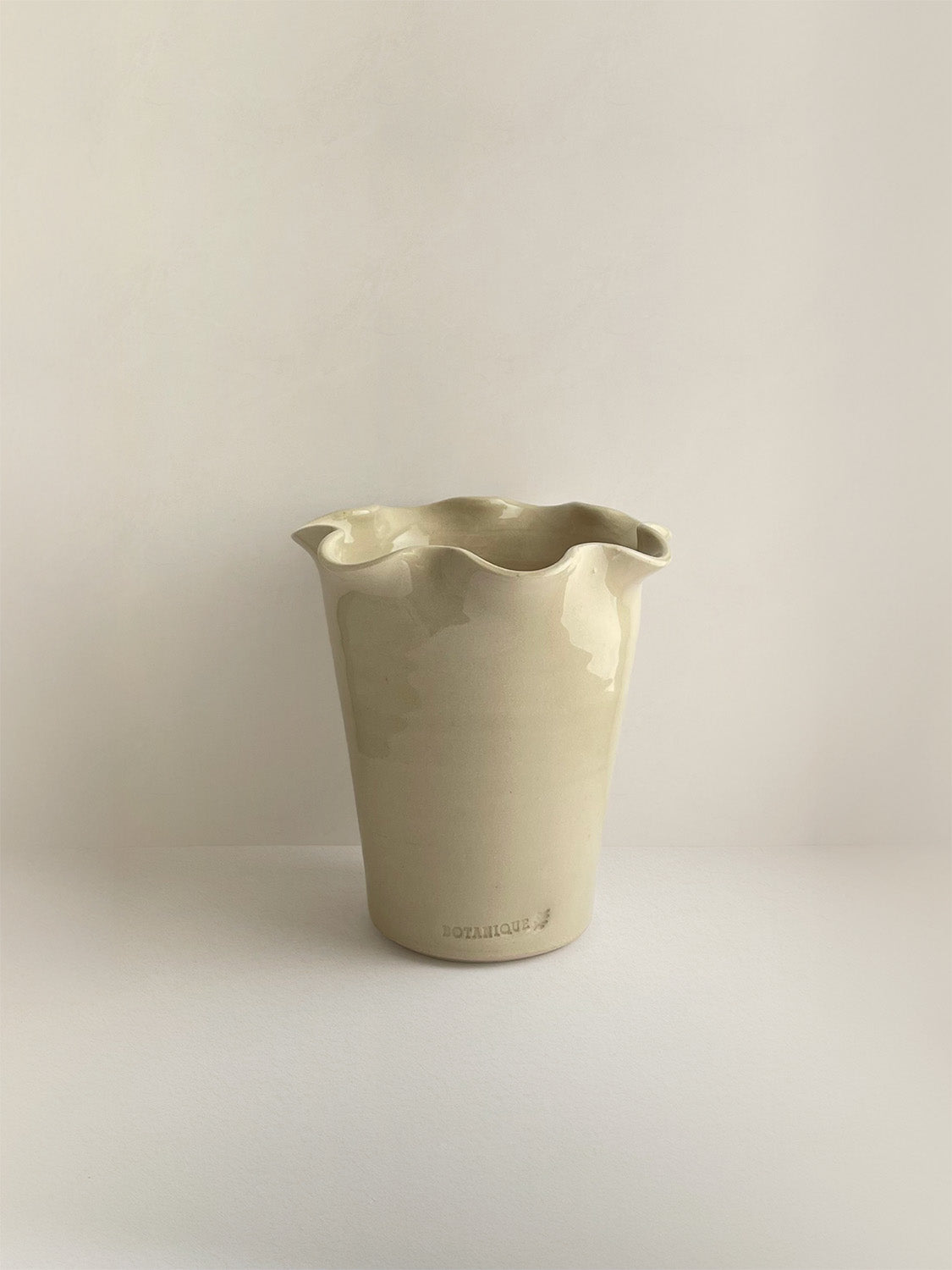 A handmade ceramic cream coloured vase with a frilled, wavy rim and maker's stamp on a neutral background.