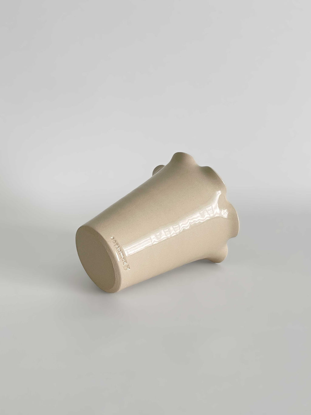 A handmade ceramic vase with a flared, frilled rim and a smooth gloss finish in a light beige colour, lying on its side set against a neutral light grey background.