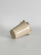 A handmade ceramic vase with a flared, frilled rim and a smooth gloss finish in a light beige colour, lying on its side set against a neutral light grey background.