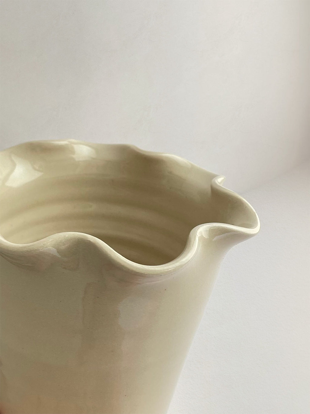 Close-up of a handmade ceramic cream coloured vase with a frilled, wavy rim and gloss finish on a neutral background.