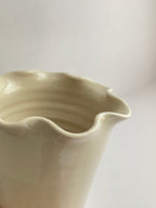 Close-up of a handmade ceramic cream coloured vase with a frilled, wavy rim and gloss finish on a neutral background.