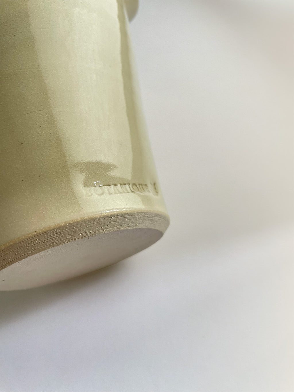 Close-up of a handmade ceramic cream coloured vase with maker's stamp and an unglazed base on a neutral background.