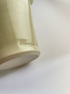 Close-up of a handmade ceramic cream coloured vase with maker's stamp and an unglazed base on a neutral background.