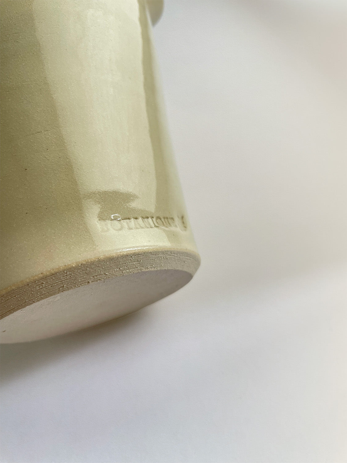 Close-up of a handmade ceramic cream coloured vase with maker's stamp and an unglazed base on a neutral background.