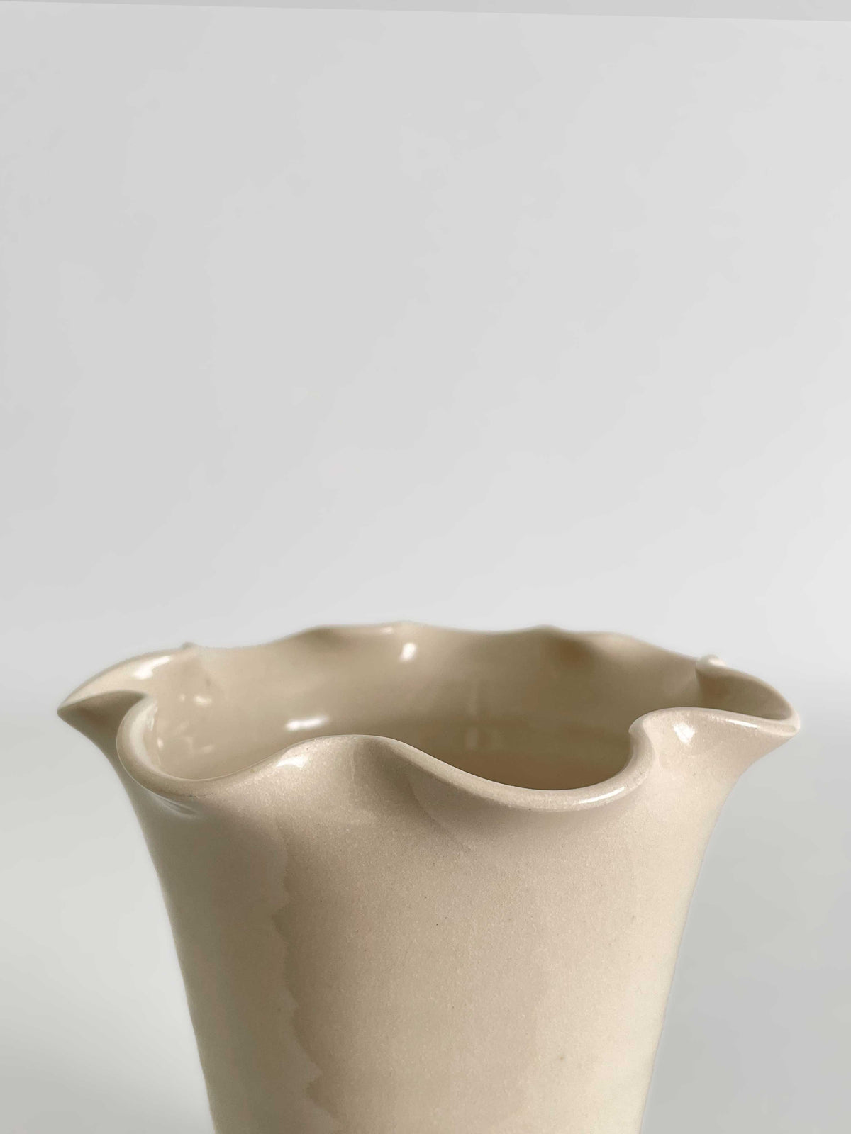 Close-up of a handmade ceramic vase with a flared, frilled rim and a smooth gloss finish in a light beige colour, set against a neutral light grey background.
