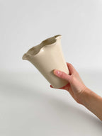 A hand holding a handmade ceramic vase with a flared, frilled rim and a smooth gloss finish in a light beige colour, set against a neutral light grey background.