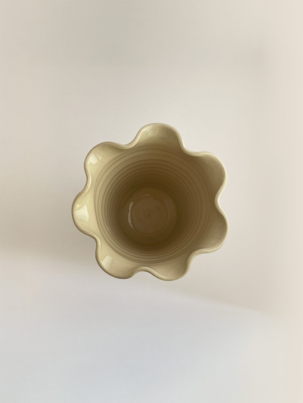 A handmade ceramic cream coloured vase with a frilled, wavy rim, on a neutral background viewed from above.