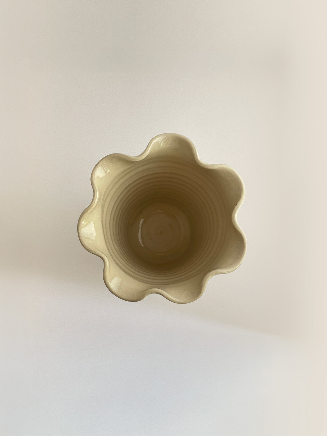 A handmade ceramic cream coloured vase with a frilled, wavy rim, on a neutral background viewed from above.