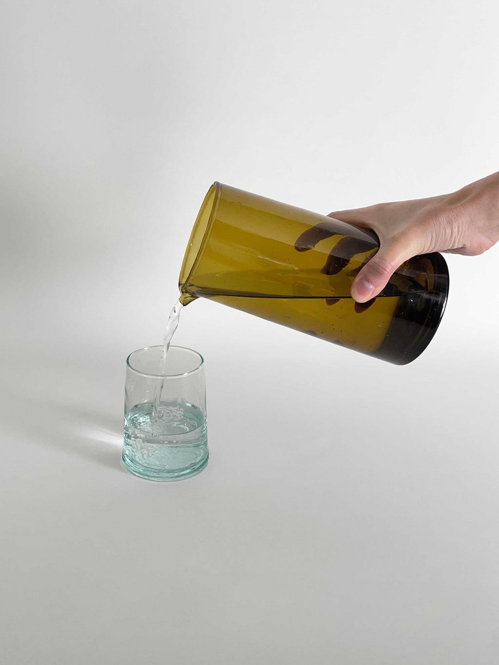 A hand pouring water from an amber glass carafe into a clear tumbler made from recycled glass with a light aqua tint, set against a light neutral background.