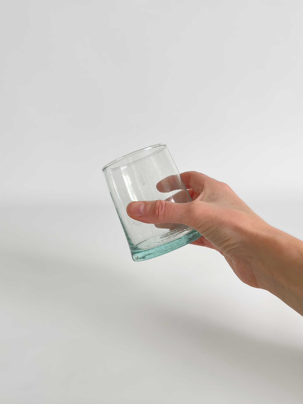 A hand holding a handmade clear tumbler made from recycled glass with a light aqua tint, on a light neutral background.