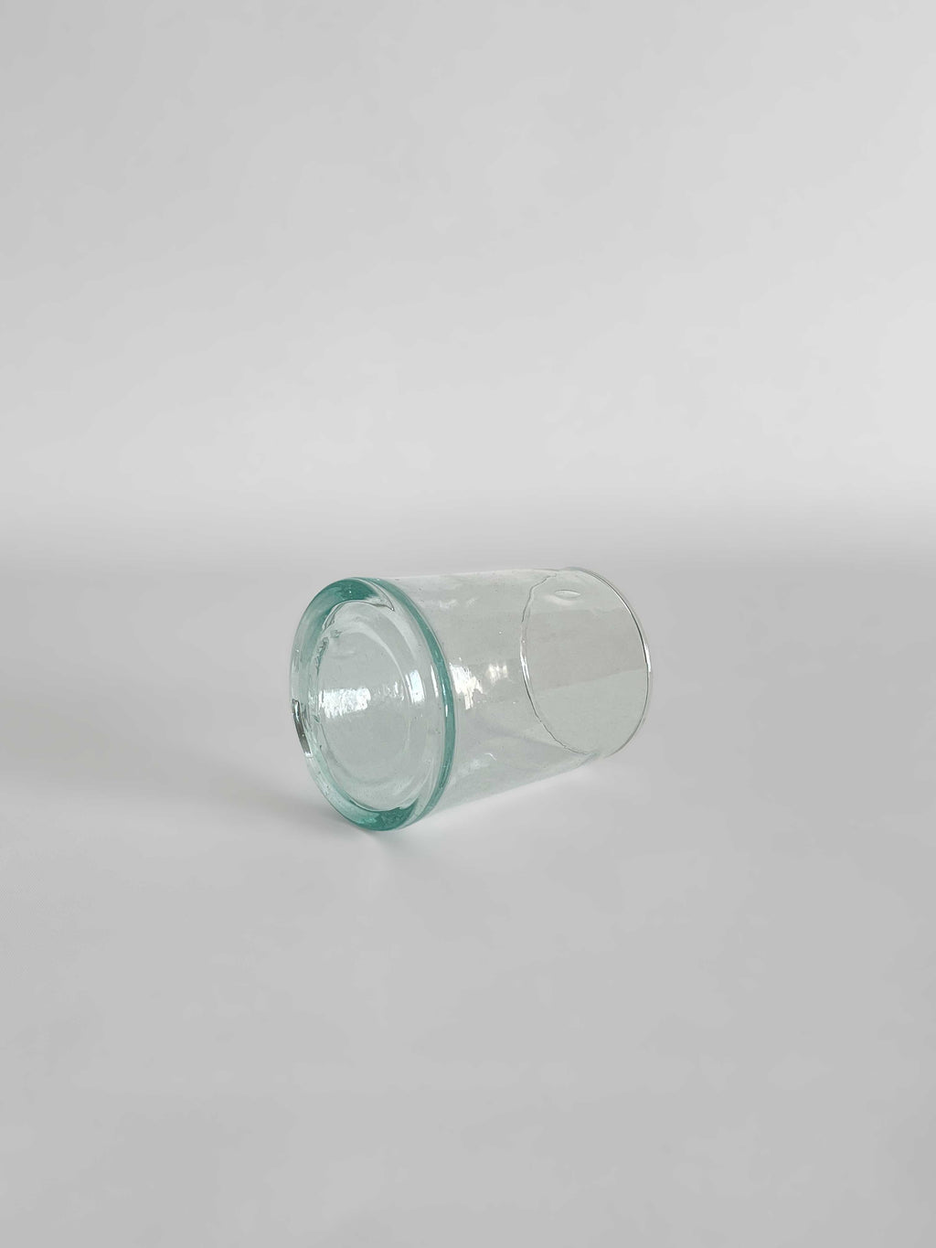 A handmade clear tumbler made from recycled glass with a light aqua tint, lying on its side on a light neutral background.