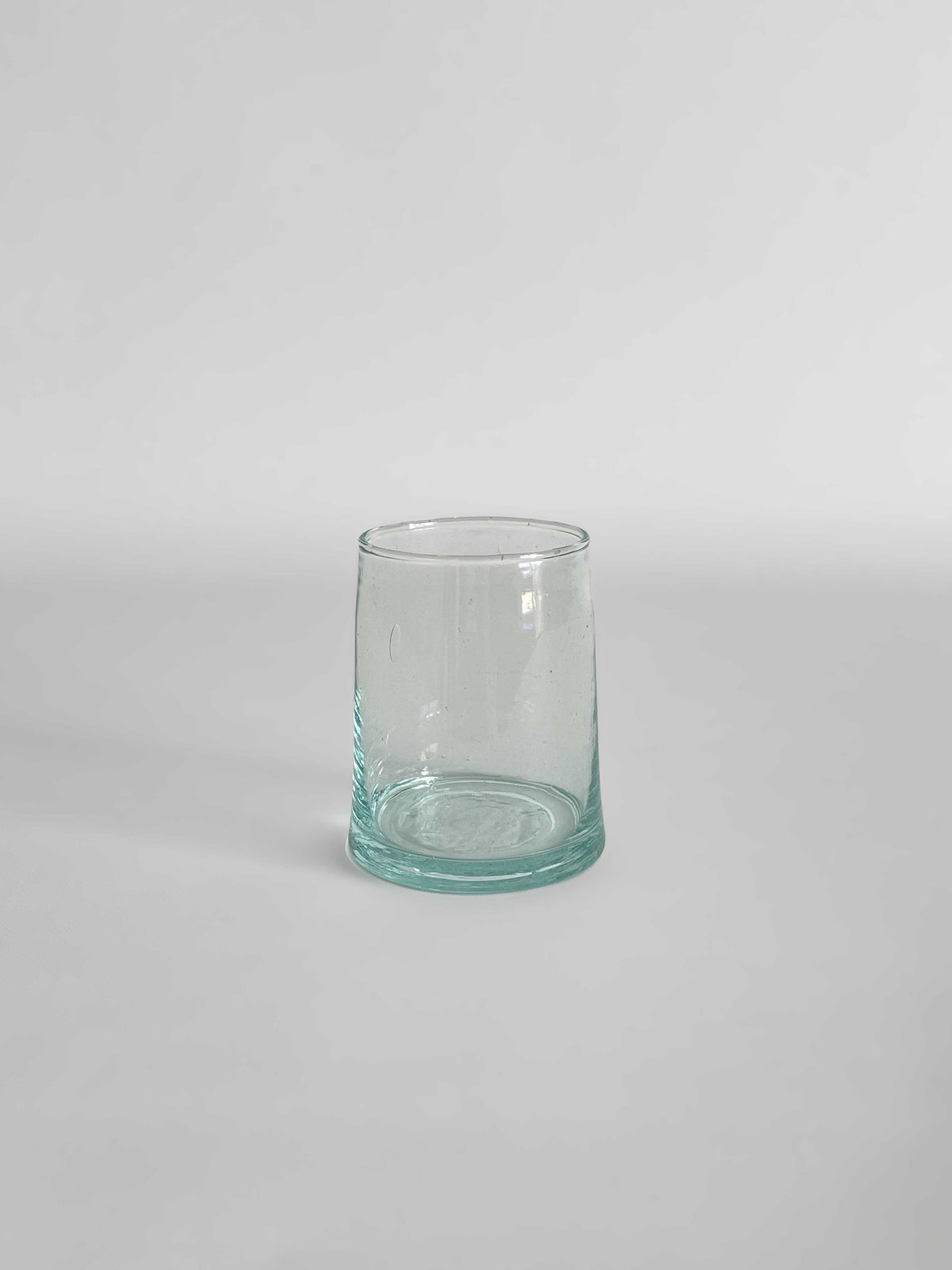 A handmade clear tumbler made from recycled glass with a light aqua tint, on a light neutral background.