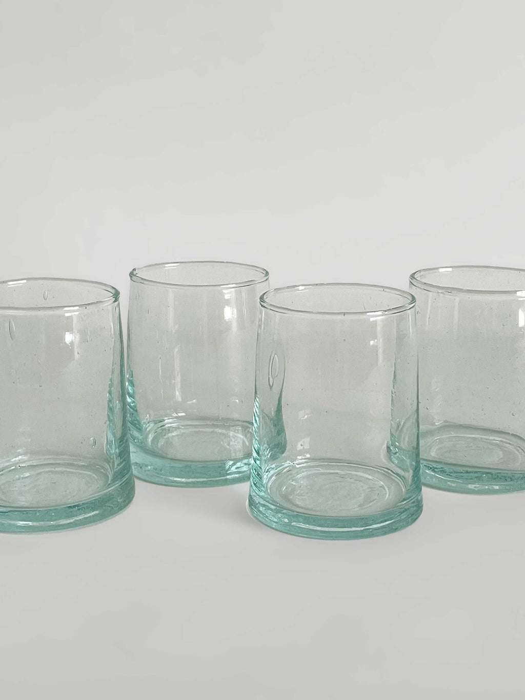 Close-up of set of four handmade clear tumblers made from recycled glass with a light aqua tint, arranged in a row on a light neutral background.