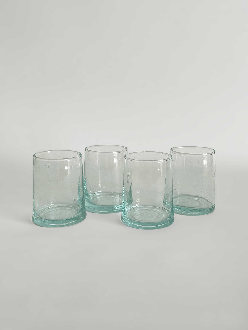 Set of four handmade clear tumblers made from recycled glass with a light aqua tint, arranged in a row on a light neutral background.