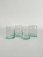 Set of four handmade clear tumblers made from recycled glass with a light aqua tint, arranged in a row on a light neutral background.