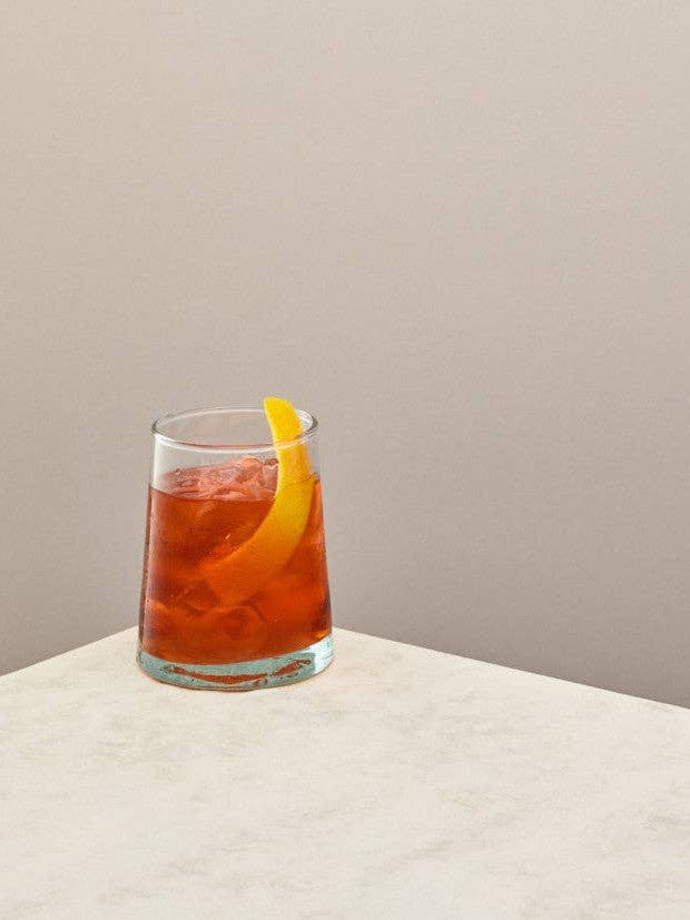 A single handmade recycled glass tumbler filled with a red drink and lemon twist on a light surface.