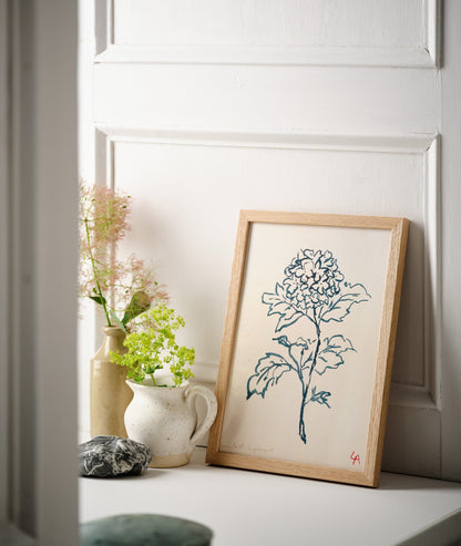 Art print of a Hydrangea flower in teal ink on vintage paper in a natural oak frame, sitting on a white painted windowsill alongside two stoneware vessels with wildflowers and a large dark grey pebble