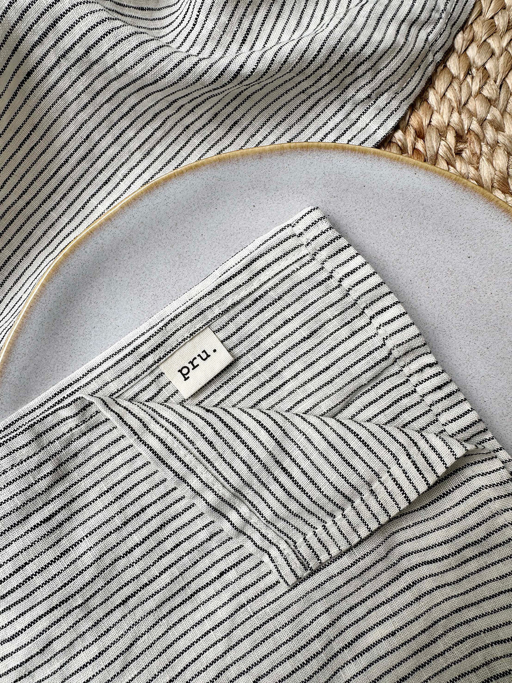 100% linen napkin set in black and ivory stripe with 'pru' label, on a white plate with woven placemat.