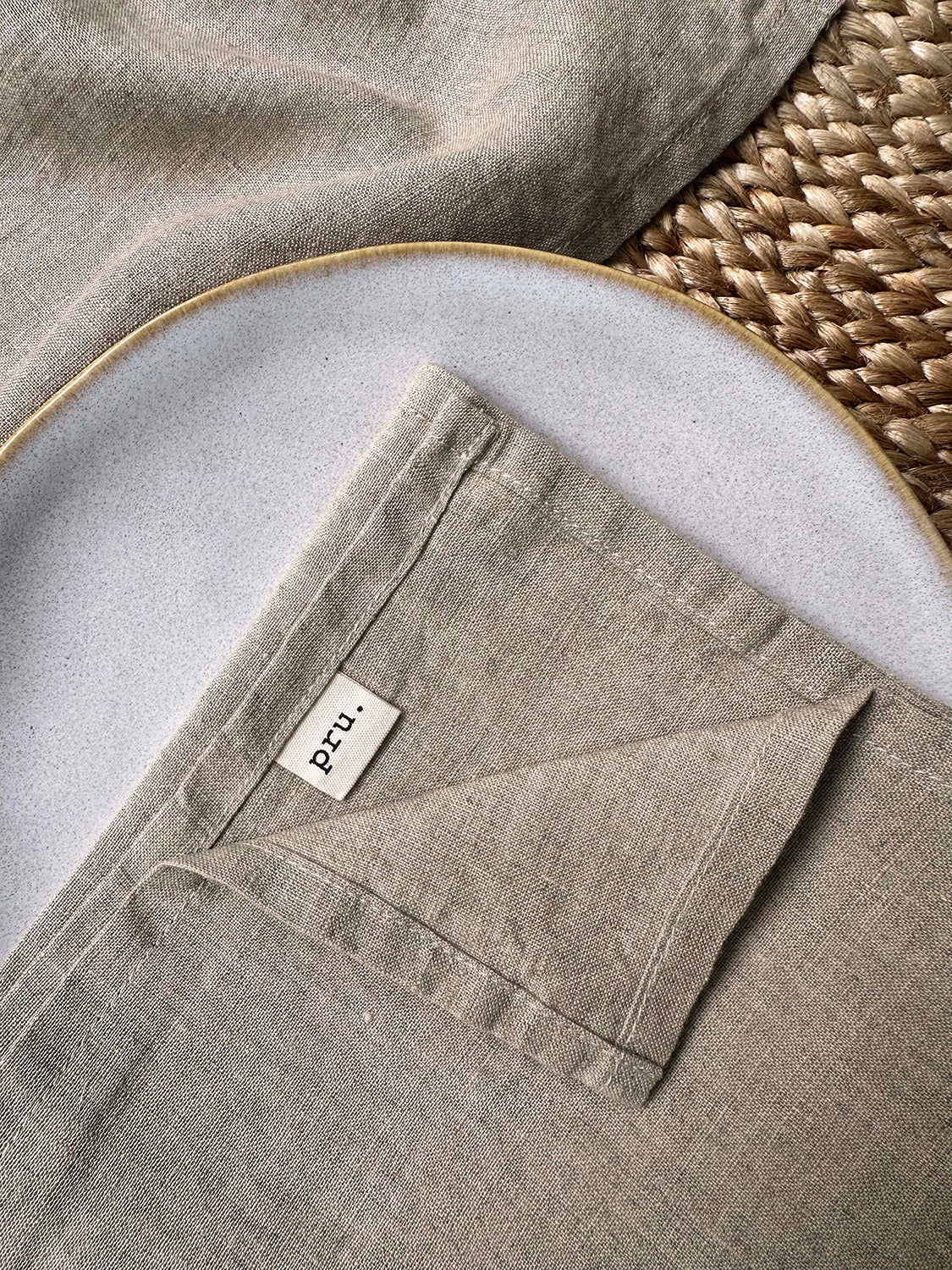 100% linen napkin set in natural colour with 'pru' label, on a white plate with woven placemat.