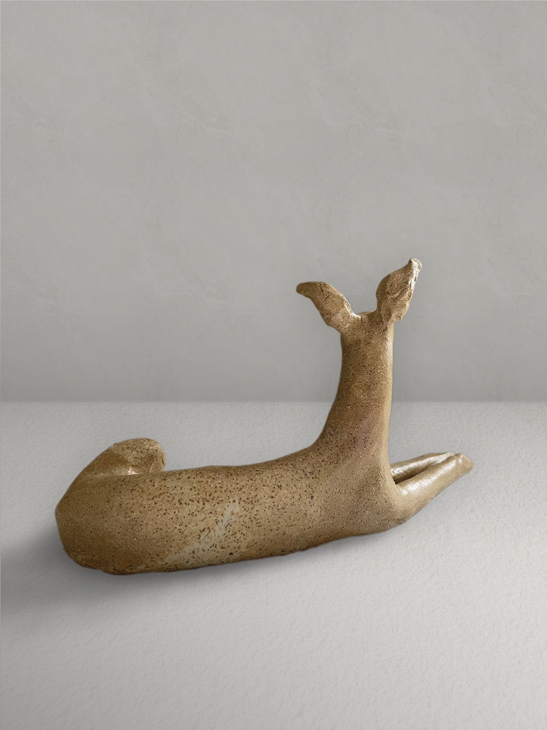 Lounging stoneware sighthound sculpture with a deep golden glaze, on a light taupe surface with a textured taupe wall behind - back view.