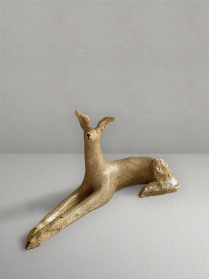 Lounging stoneware sighthound sculpture with a deep golden glaze, on a light taupe surface with a textured taupe wall behind - front view.