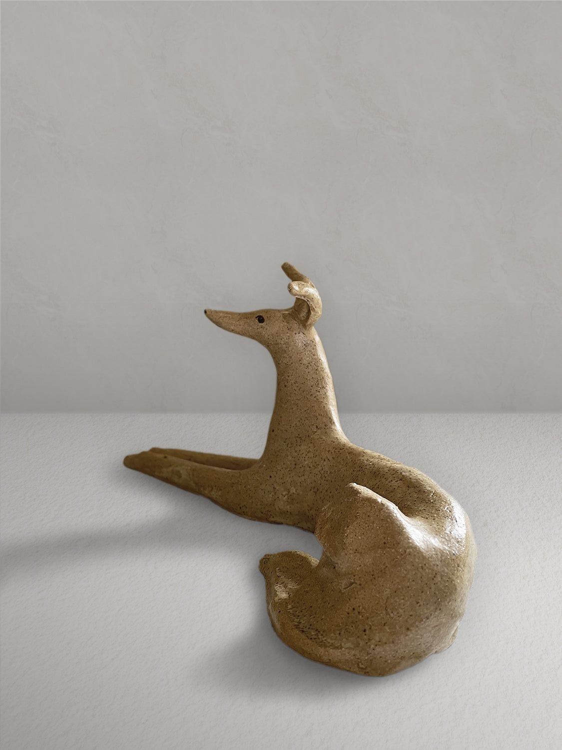 Lounging stoneware sighthound sculpture with a deep golden glaze, on a light taupe surface with a textured taupe wall behind - left side view.