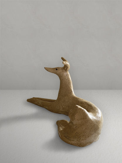 Lounging stoneware sighthound sculpture with a deep golden glaze, on a light taupe surface with a textured taupe wall behind - left side view.