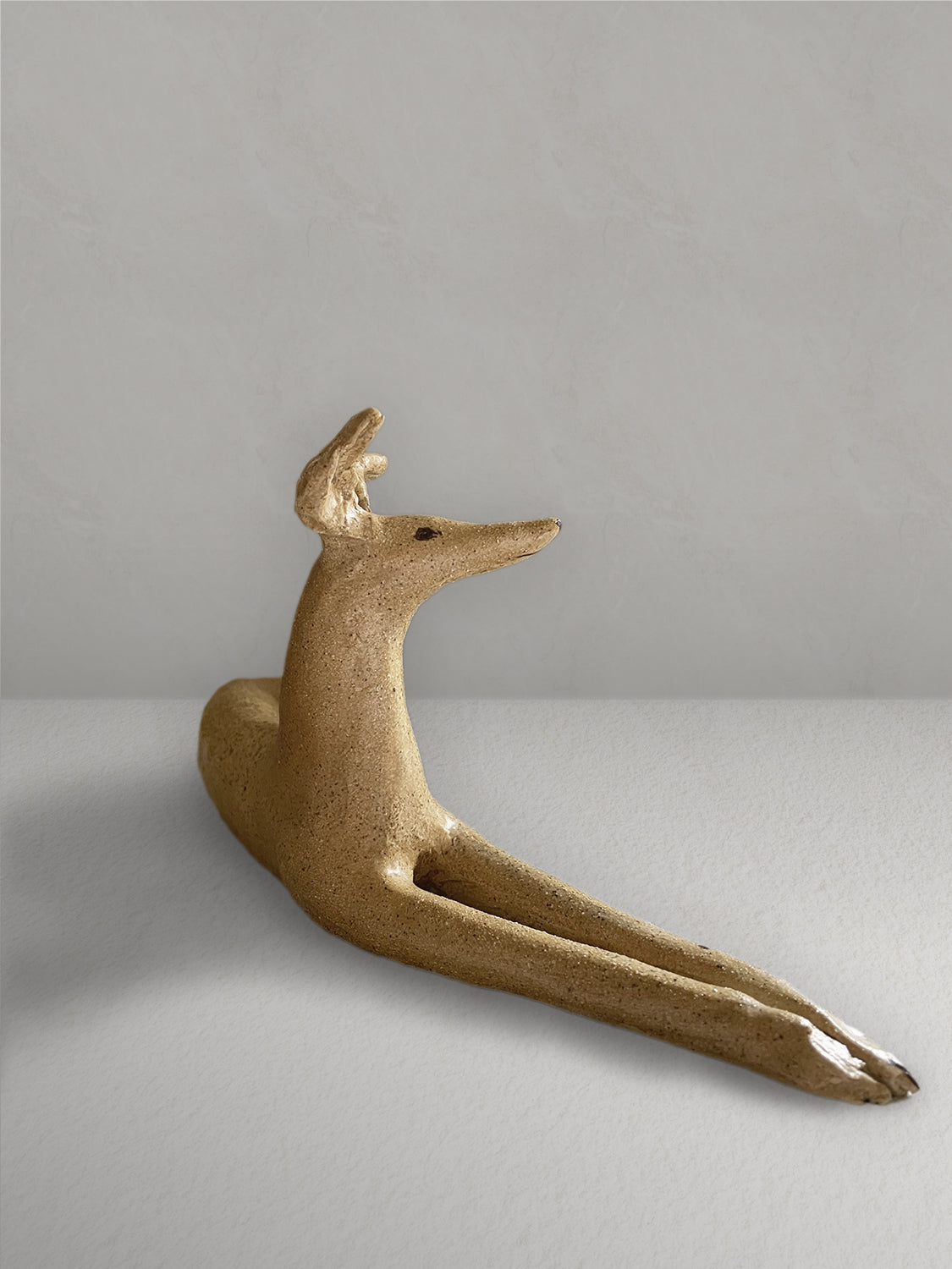 Lounging stoneware sighthound sculpture with a deep golden glaze, on a light taupe surface with a textured taupe wall behind - right side view.