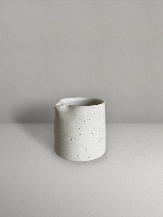 Ceramic small milk jug in glossy off-white speckled glaze on a light taupe surface with a textured taupe wall behind - front view.