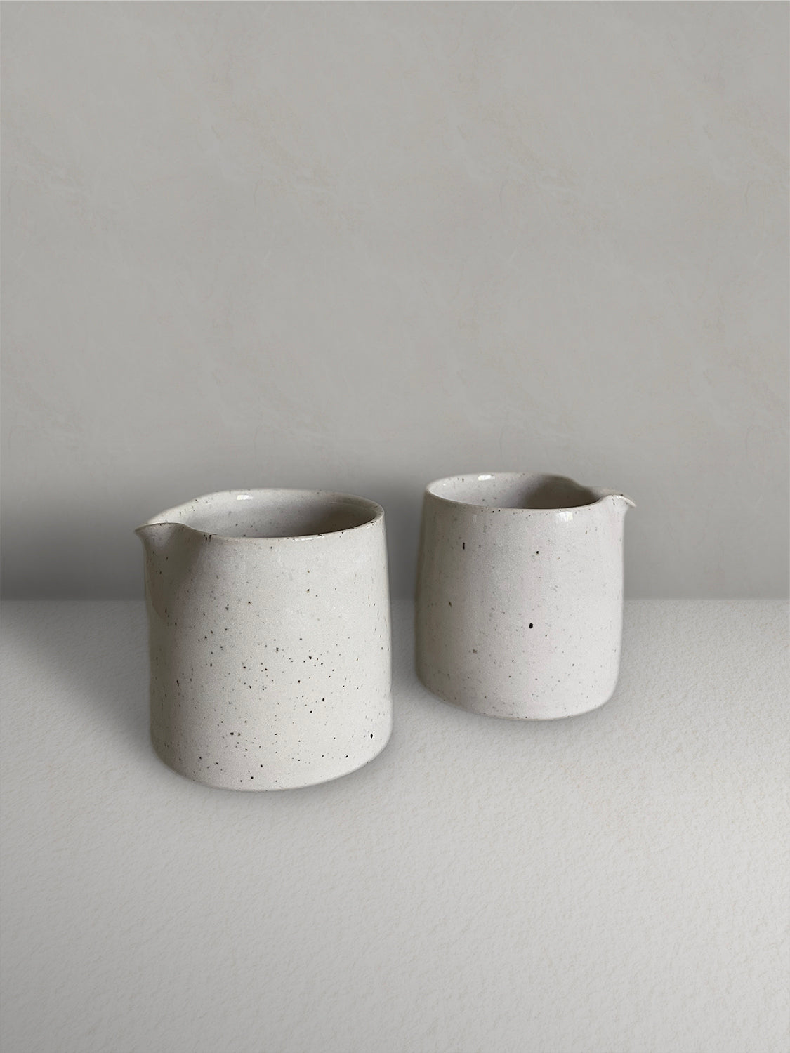 Pair of ceramic small milk jugs in glossy off-white speckled glaze facing opposite directions on a light taupe surface with a textured taupe wall behind - front view.