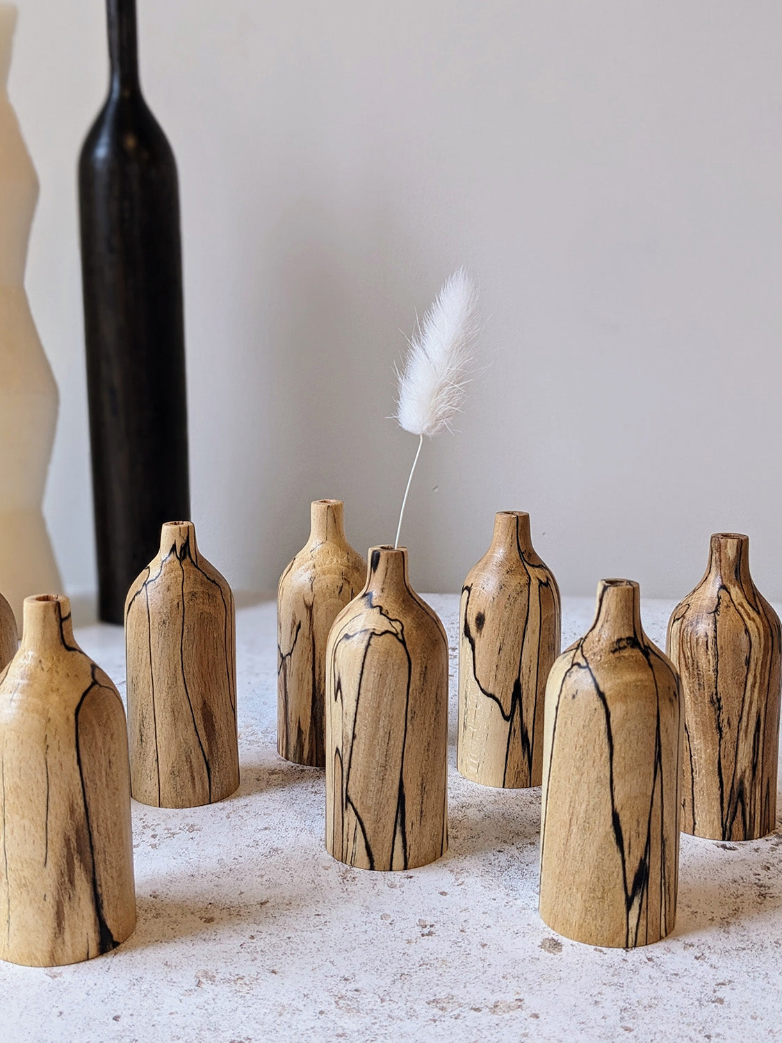 Group of seven miniature bud vases in spalted Beech with tall vase in dark stained timber in the background, on a mottled off-white surface against a plain taupe wall - front view.