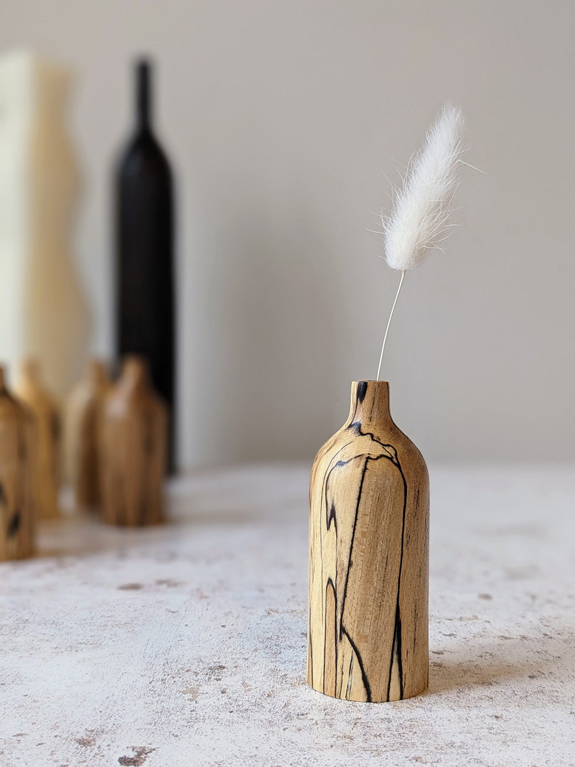 Miniature bud vase in spalted Beech with dried stem, on a mottled off-white surface against a plain taupe wall - front view.