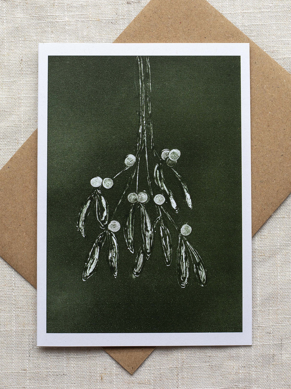Botanical Christmas card with mistletoe design on a dark green background, placed on a linen surface with a Kraft paper envelope.