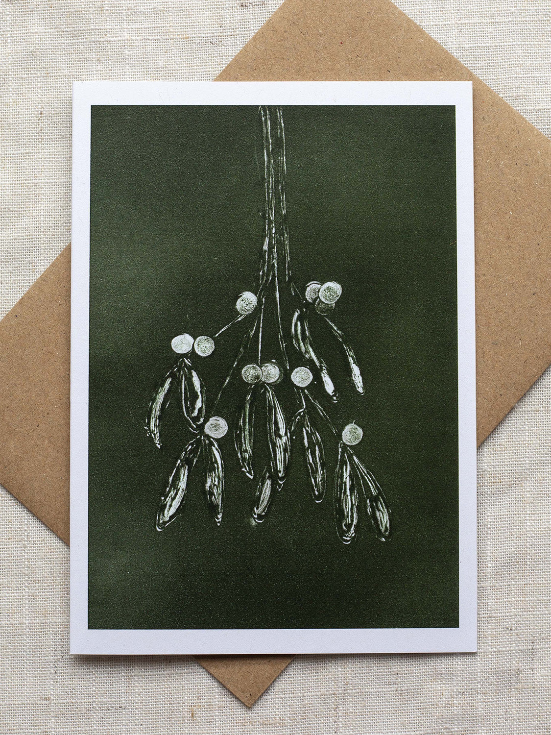 Botanical Christmas card with mistletoe design on a dark green background, placed on a linen surface with a Kraft paper envelope.